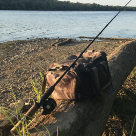 A fishing rod and duffel bag rest on a log on the shore of a lake.