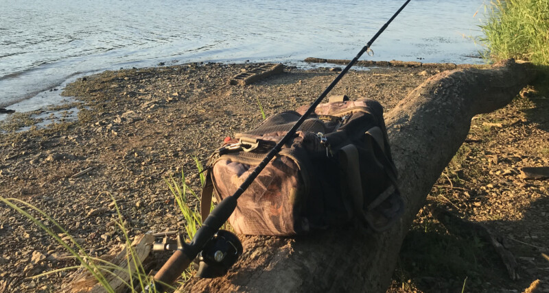 A fishing rod and duffel bag rest on a log on the shore of a lake.