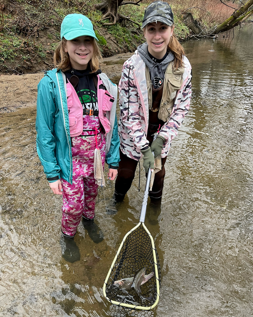 Two young anglers stand in shallow water. One of the anglers holds a net containing two trout in the water.