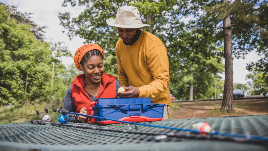 Two anglers look through a tackle box at a picnic table. Their fishing rods are also laying on the table.