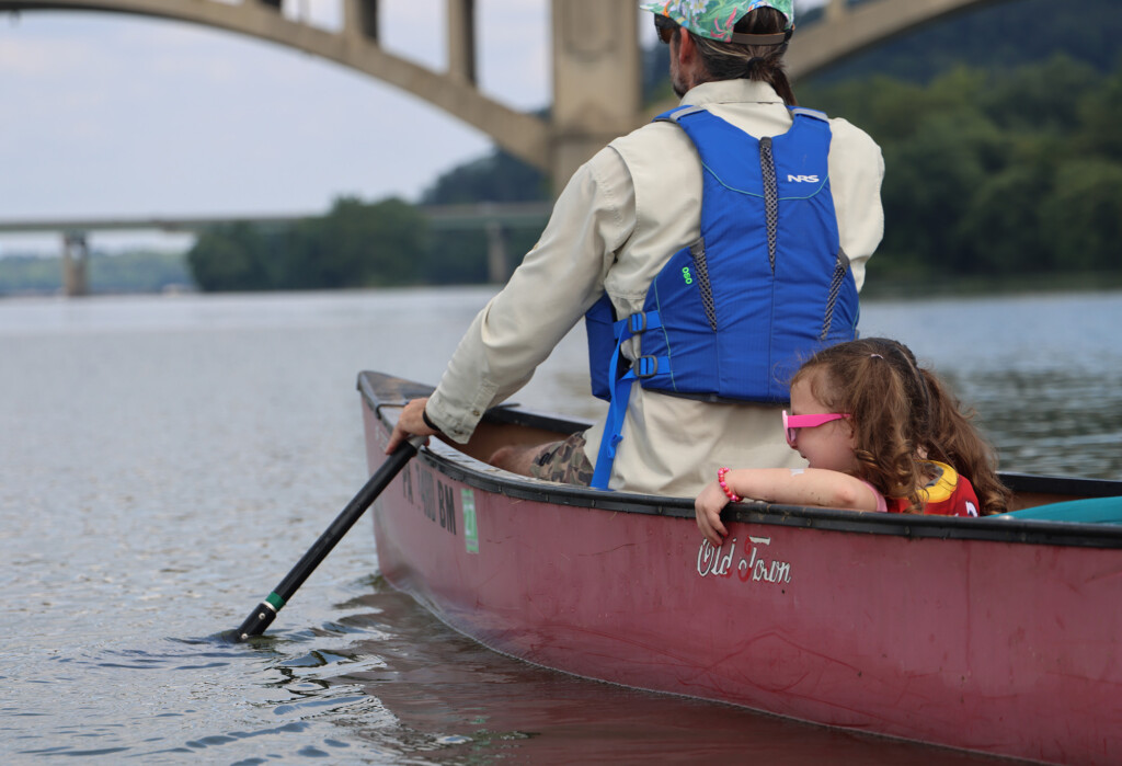 Man wearing a life jacket paddling the bow of a canoe with a young girl seated behind him wearing a life jacket and sunglasses.