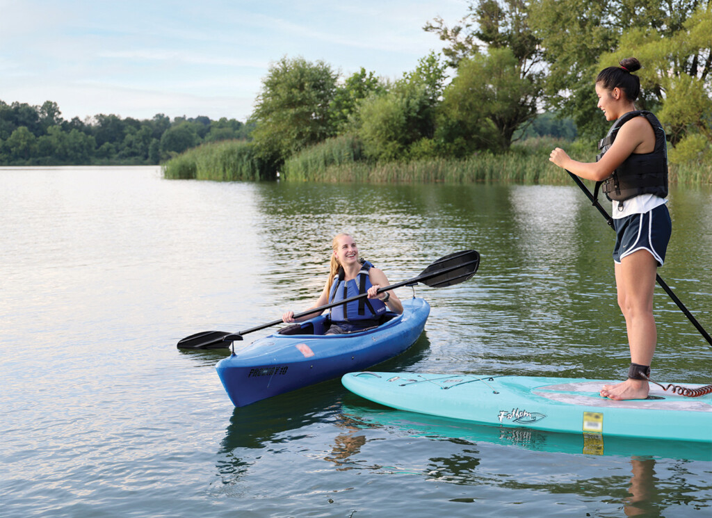 A woman wearing a life jacket holding a paddle sitting in a kayak next to a woman wearing a life jacket holding a paddle while standing on a paddleboard on Marsh Creek Lake in PA.