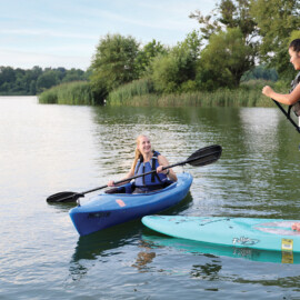 A woman wearing a life jacket holding a paddle sitting in a kayak next to a woman wearing a life jacket holding a paddle while standing on a paddleboard on Marsh Creek Lake in PA.