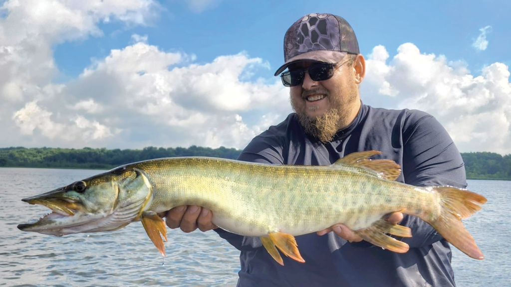 An angler holds a Muskellunge with a lake in the background.