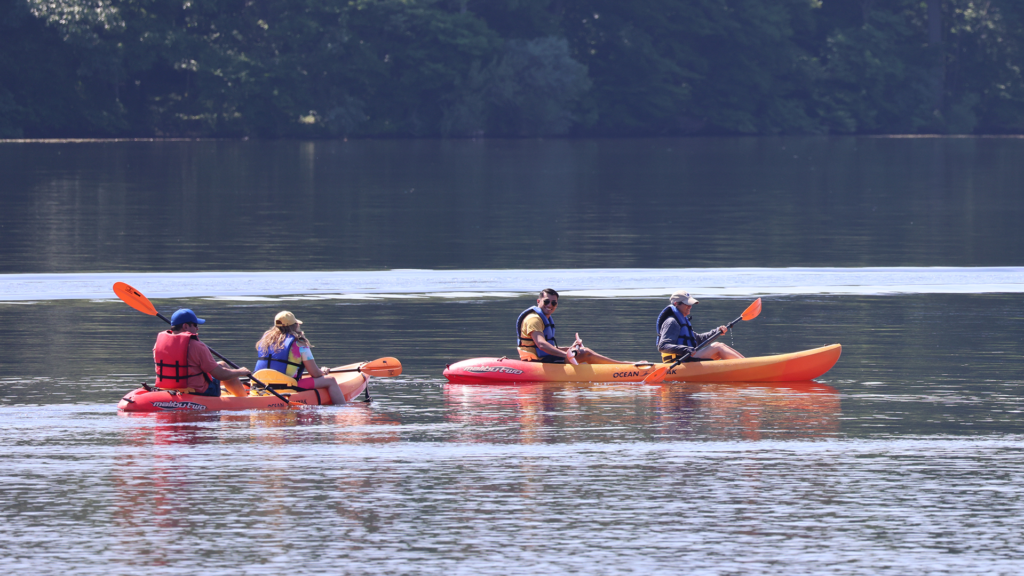 Four people paddle in two tandem kayaks on a lake. All four are wearing life jackets.