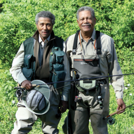 Two anglers pose for a photo. Both men are wearing waders and other fishing ger and holding fishing rods in their hands.