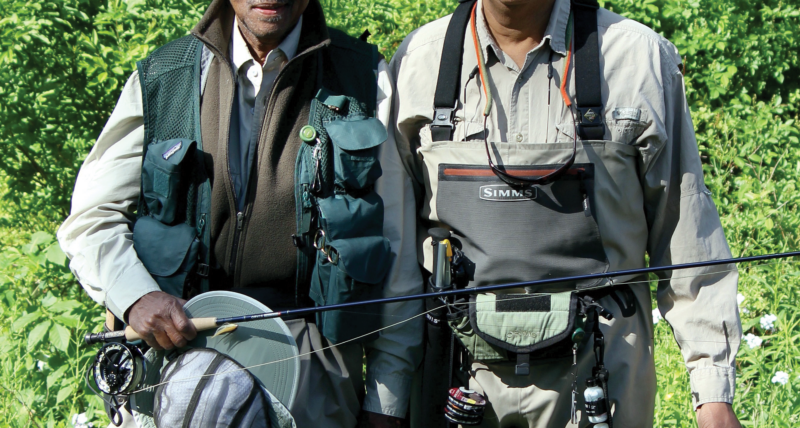 Two anglers pose for a photo. Both men are wearing waders and other fishing ger and holding fishing rods in their hands.