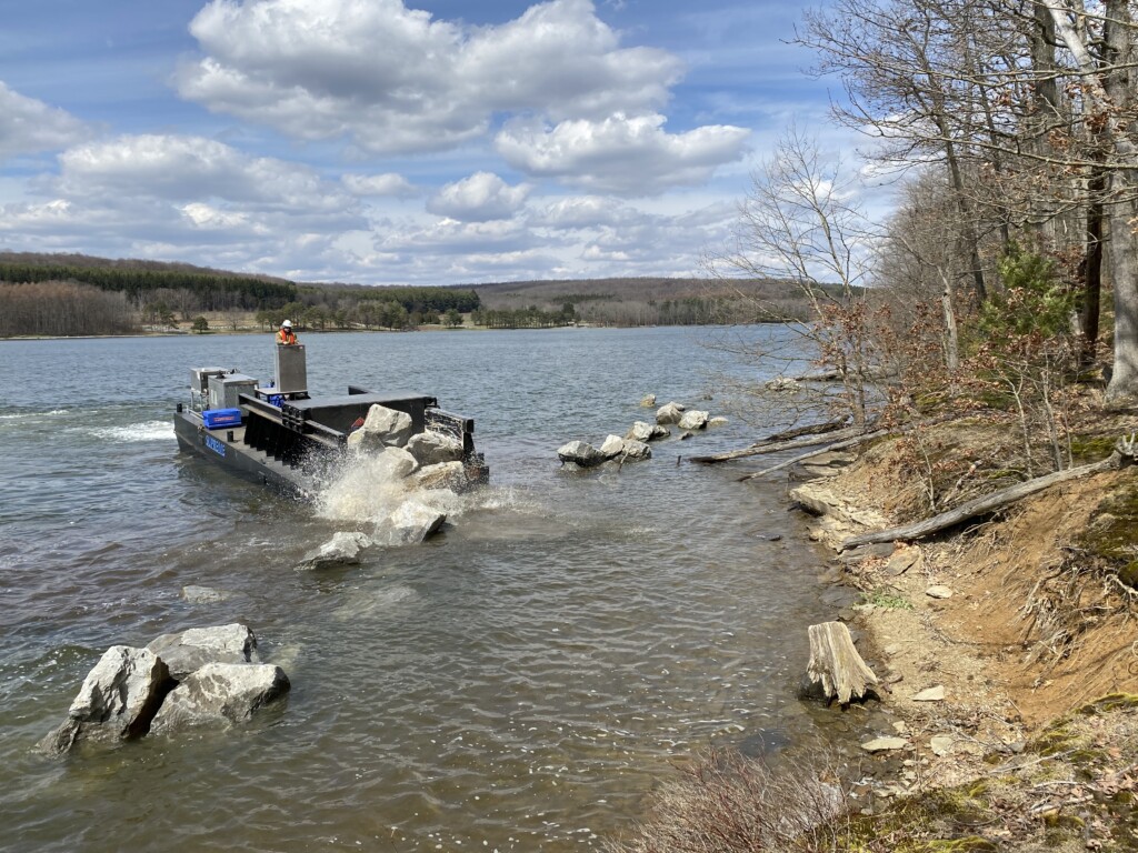 A barge drops large rocks into the water along an eroded lake shore. One person operates the barge.