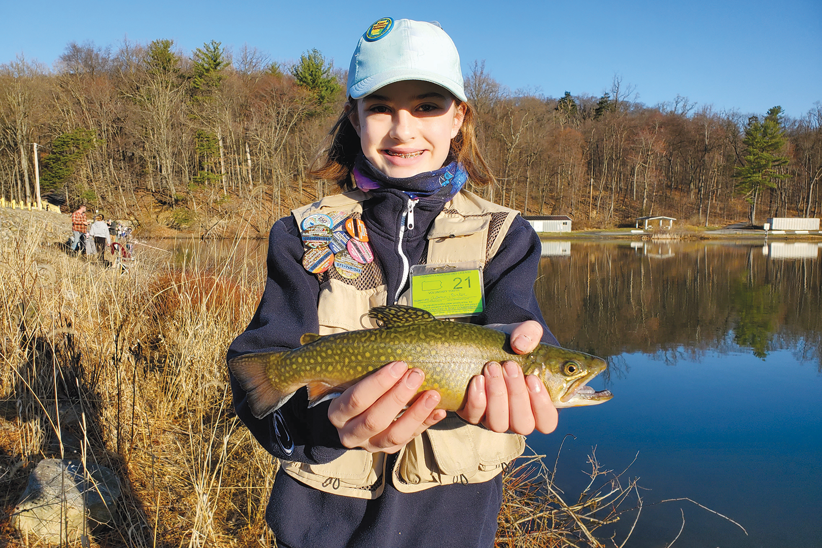 A young angler poses for a photo while holding a trout. She's standing on a lakeshore and wearing fishing gear.