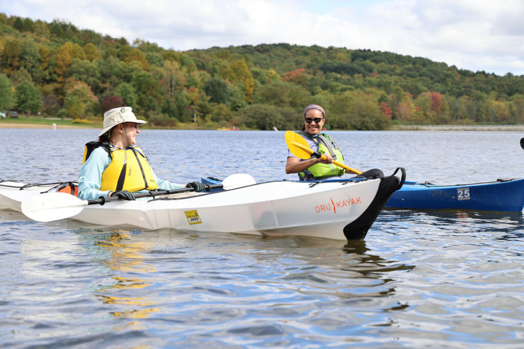 Woman wearing a life jacket sitting in a portable kayak with a launch permit. And another woman wearing sunglasses and a life jacket sitting in a blue registered kayak.