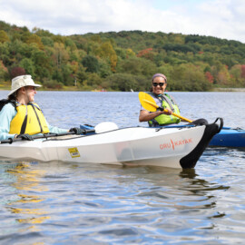 Woman wearing a life jacket sitting in a portable kayak with a launch permit. And another woman wearing sunglasses and a life jacket sitting in a blue registered kayak.