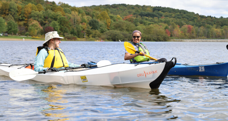 Woman wearing a life jacket sitting in a portable kayak with a launch permit. And another woman wearing sunglasses and a life jacket sitting in a blue registered kayak.