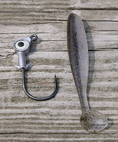 A Large Round Head Jig with an eye at the top of the hook sitting next to a silver Paddle Tail lure for imitating minnnows.