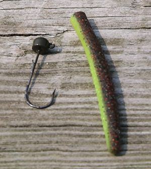 A Ned Rig Jig with a weighted top on a hook sitting next to a straight plastic lure resembling a small minnow used for luring in bass.