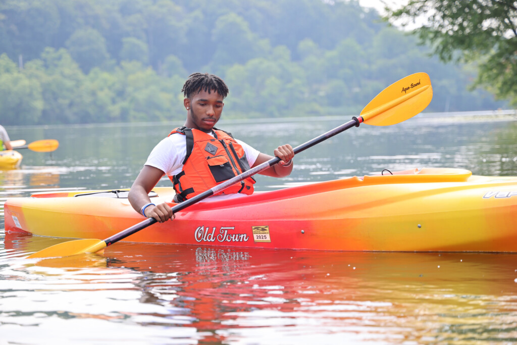 Medium shot of young man on lake in a bright yellow, red and orange kayak. He is holding the paddle in two hands and he's wearing a life jacket. 