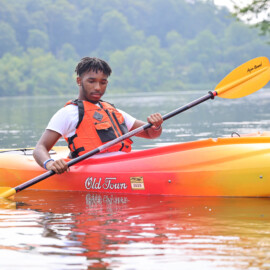 Medium shot of young man in a kayak on a lake with a paddle in his hand. He's wearing a life jacket.