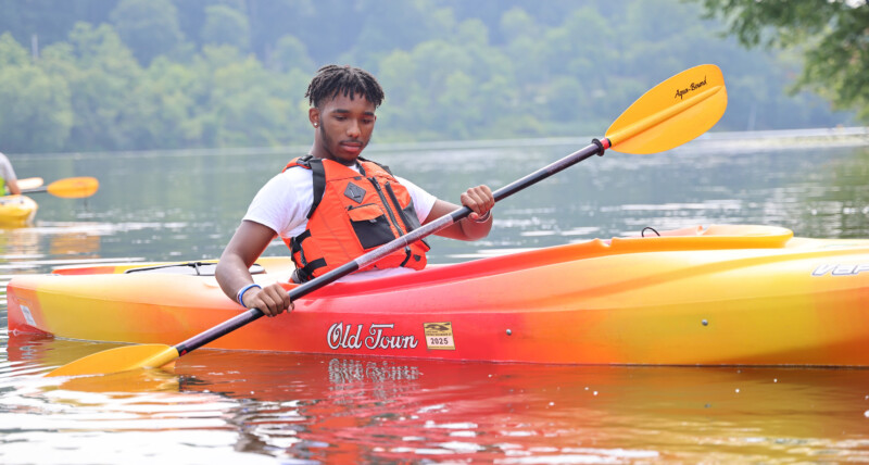 Medium shot of young man in a kayak on a lake with a paddle in his hand. He's wearing a life jacket.