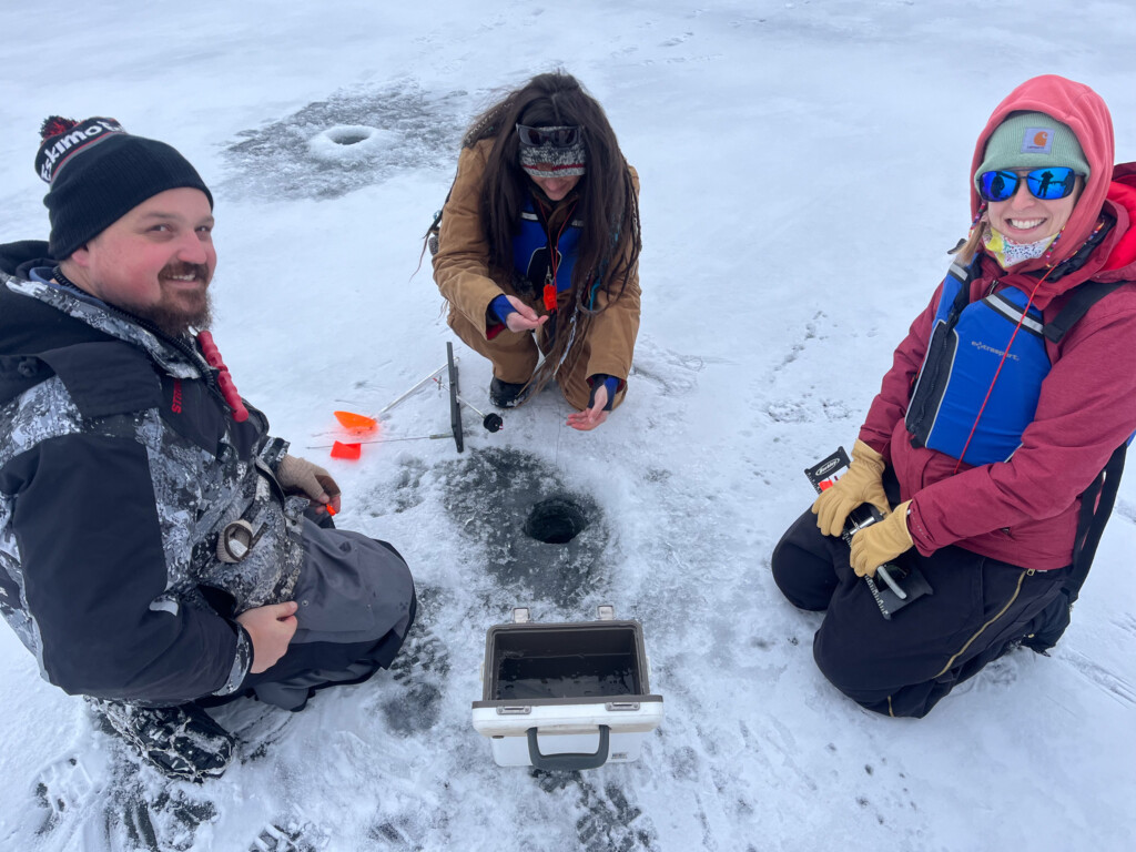 A man and two women kneeling on a frozen lake while ice fishing.