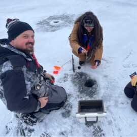 A man and woman kneeling on a frozen lake while ice fishing.