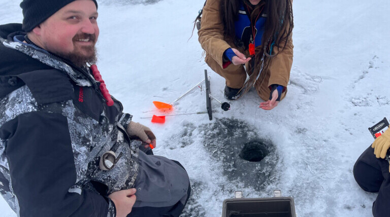 A man and woman kneeling on a frozen lake while ice fishing.