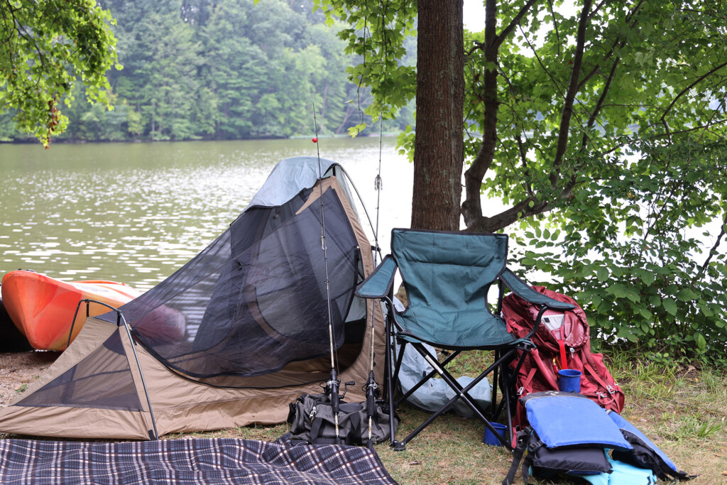 A blanket, tent, chair, backpack and kayaks set up as a campsite under some trees on the banks of a lake. 