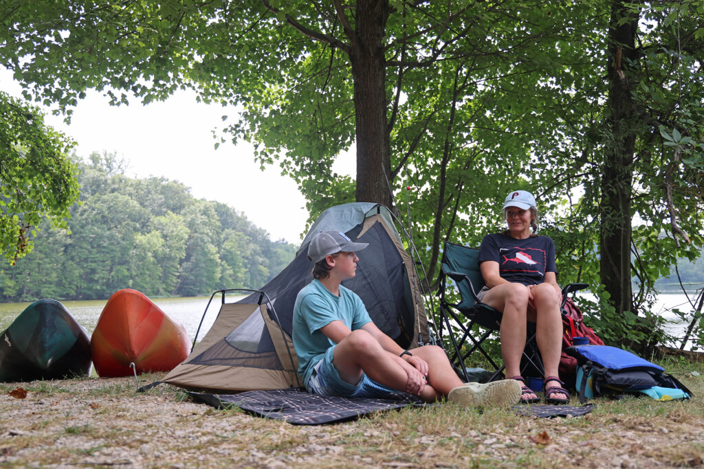 A boy and woman sitting at a camp site. Two kayaks beside their campsite. The campsite is under trees and a lake is in the background.