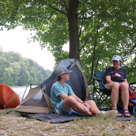 A boy and woman sitting at a camp site. Two kayaks beside their campsite. The campsite is under trees and a lake is in the background.