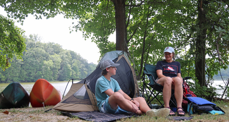 A boy and woman sitting at a camp site. Two kayaks beside their campsite. The campsite is under trees and a lake is in the background.