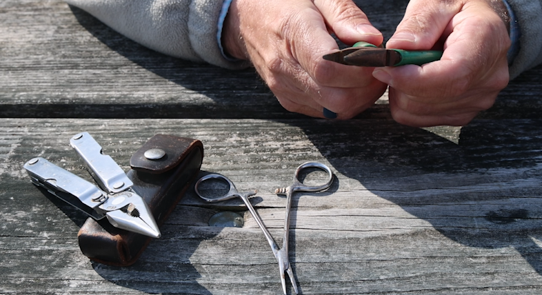 A man's hand holding needle nose pliers with forceps and a multi-tool laying on a picnic table below.