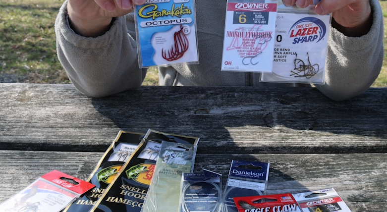 A man's hands holding bags of different sizes of baitholder hooks with a variety of other sizes laying on a picnic table below.