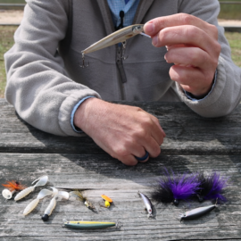 A man's hand holding a topwater lure with other artificial lures laying on a picnic table below