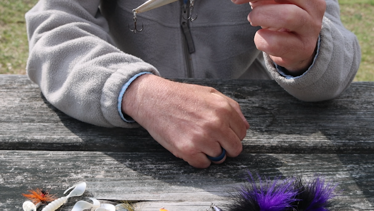 A man's hand holding a topwater lure with other artificial lures laying on a picnic table below