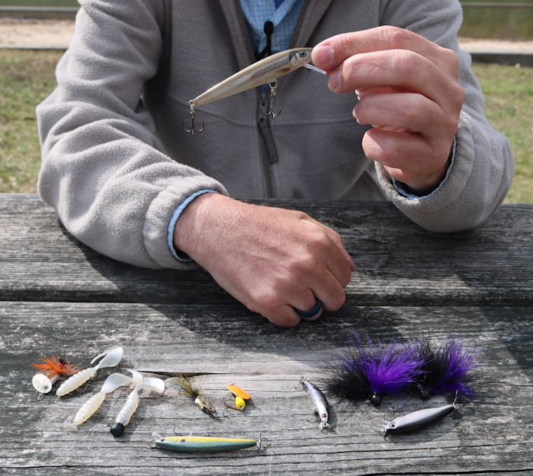 A man's hand holding a topwater lure with other artificial lures laying on a picnic table below