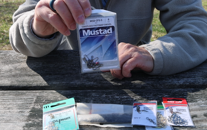 A man's hands holding a bag of snap swivels with a variety of other swivels laying on a picnic table below.