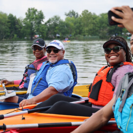 A family of four all wearing life jackets and sitting in their kayaks on a lake take a selfie with a cell phone.
