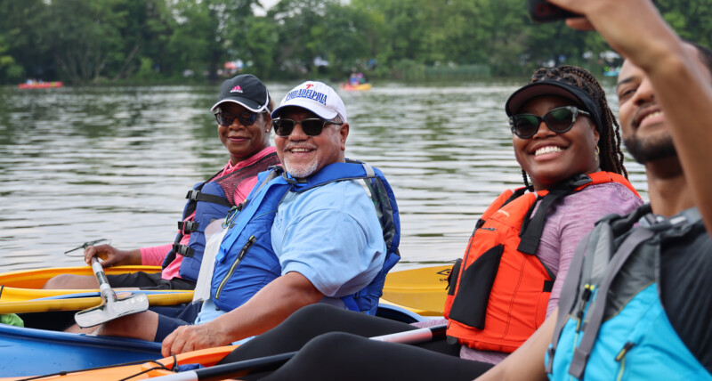 A family of four all wearing life jackets and sitting in their kayaks on a lake take a selfie with a cell phone.