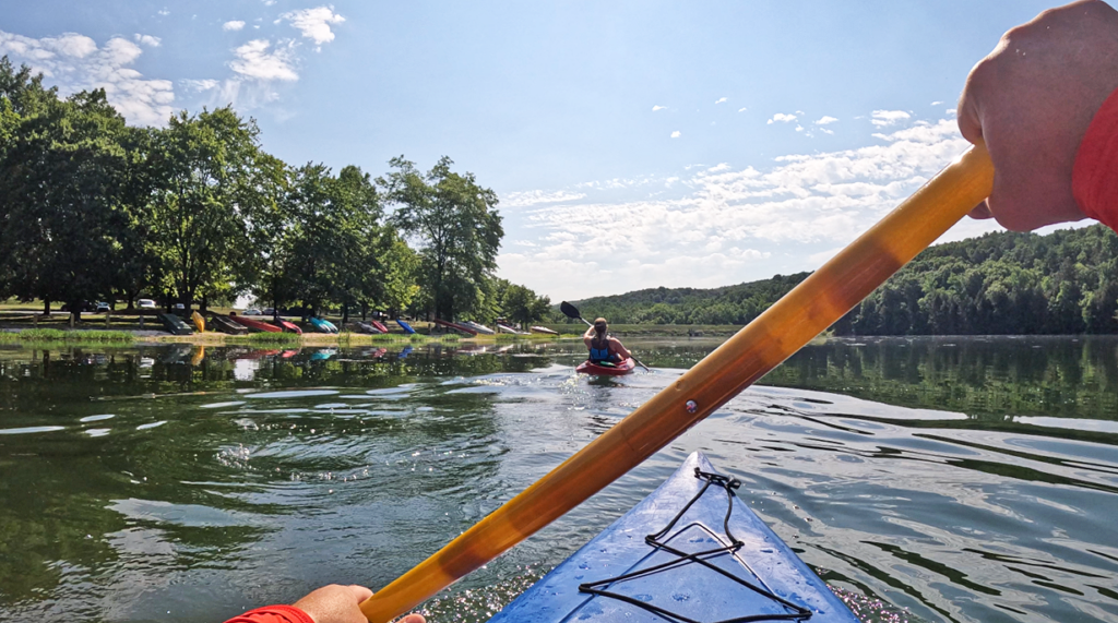 POV from a man paddling a kayak on a lake with a friend wearing a life jacket paddling in front of him.