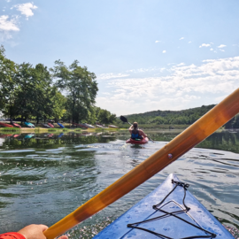 POV from a man paddling a kayak on a lake with a friend wearing a life jacket paddling in front of him.
