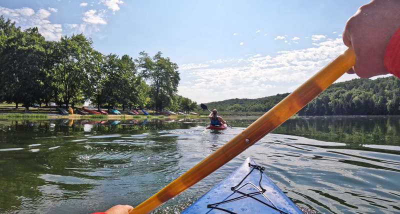 POV from a man paddling a kayak on a lake with a friend wearing a life jacket paddling in front of him.