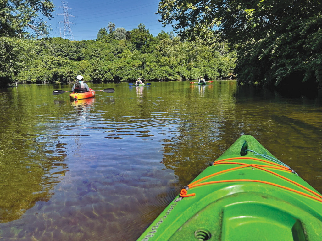 Tip of a kayak in the foreground with kayakers enjoying the calm Lehigh River and green scenery in the background.