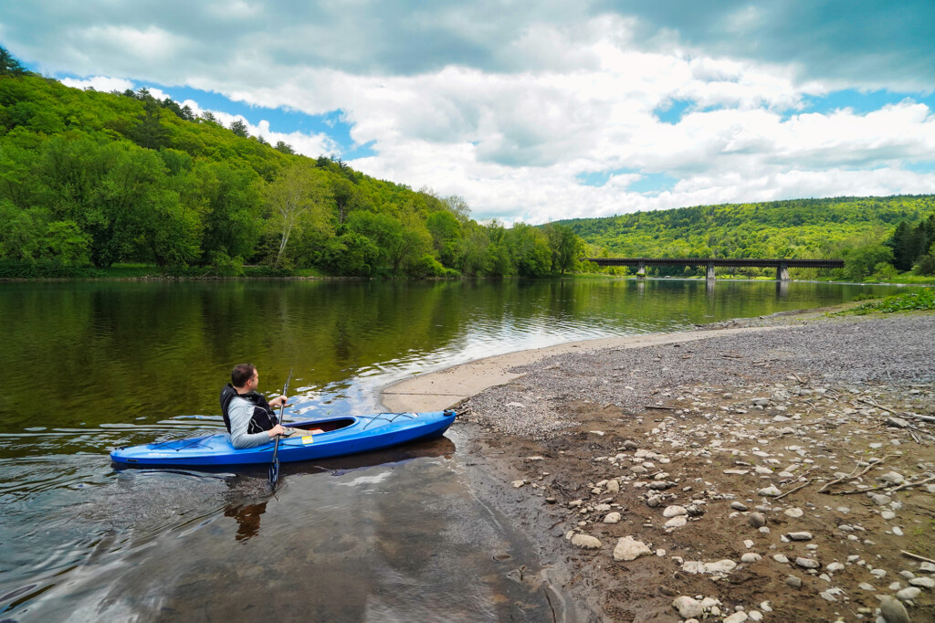 A man wearing a life jacket while sitting in a kayak along the shallow shore of the Lackawaxon River.