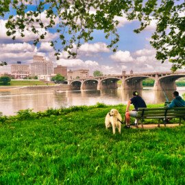 Scenic view of the riverfront in Wilkes-Barre of two men and a dog sitting on a bench while fishing.