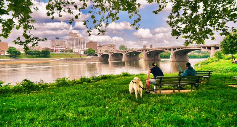 Scenic view of the riverfront in Wilkes-Barre of two men and a dog sitting on a bench while fishing.