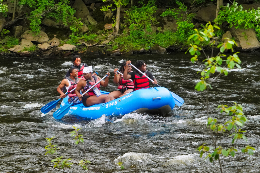 Group of young adults wearing life jackets while paddling a raft in river rapids.