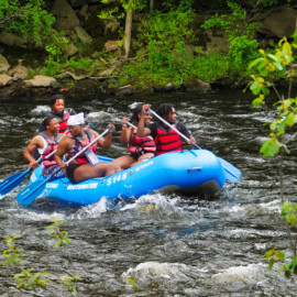 Group of young adults wearing life jackets while paddling a raft in river rapids.