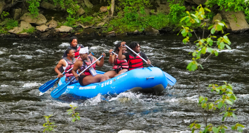 Group of young adults wearing life jackets while paddling a raft in river rapids.